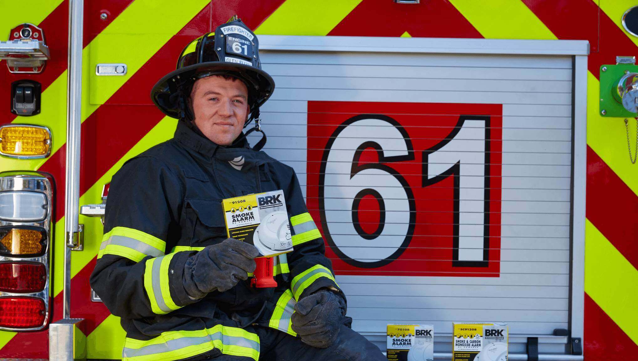 A firefighter holds a smoke alarm and stands next to a firetruck marked 61.