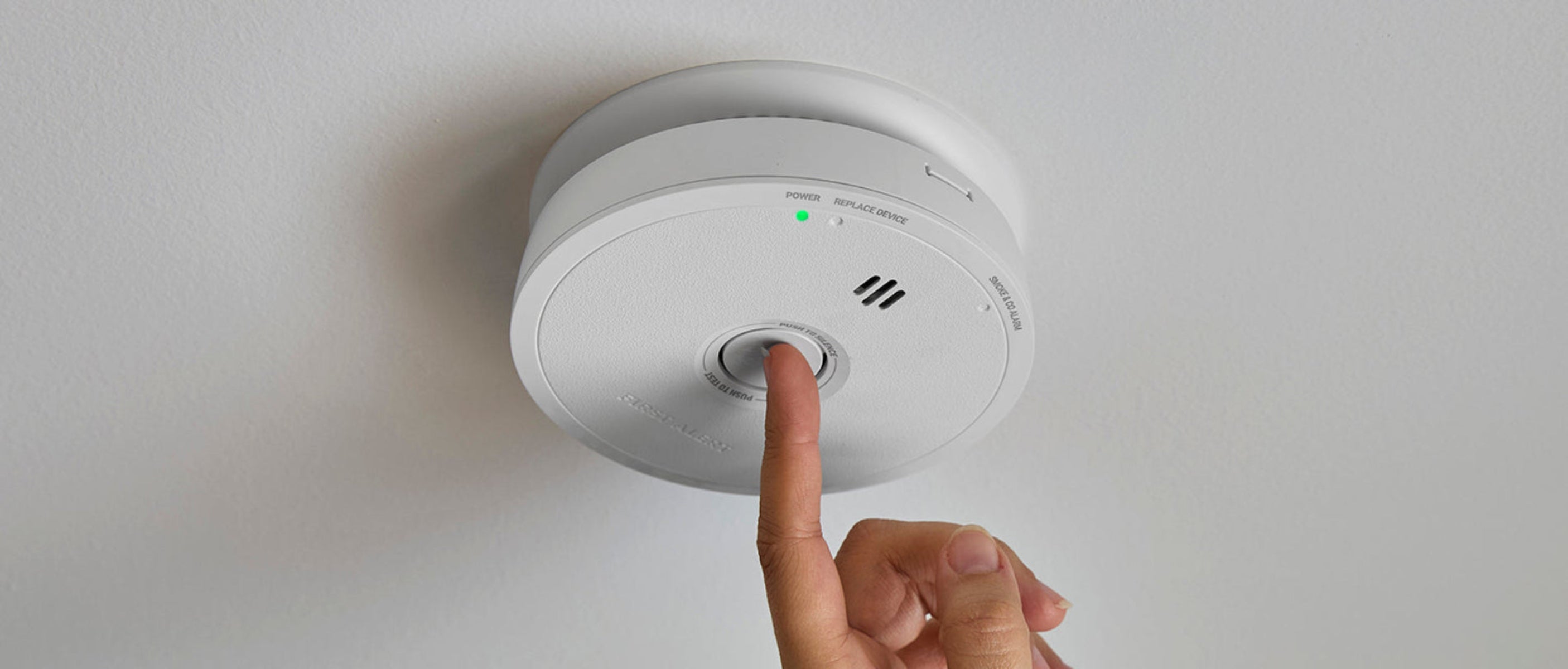 Close-up of a smoke and carbon monoxide alarm mounted on a ceiling with a hand pressing the test/reset button.