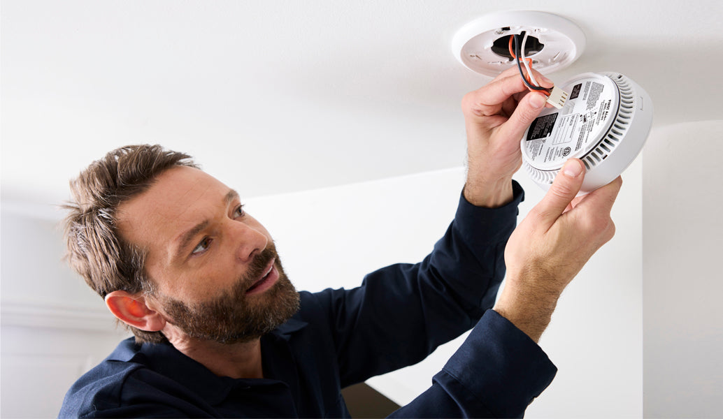 A man in a navy blue shirt installs a white round smoke detector on a white ceiling, holding the device and its attached wiring.